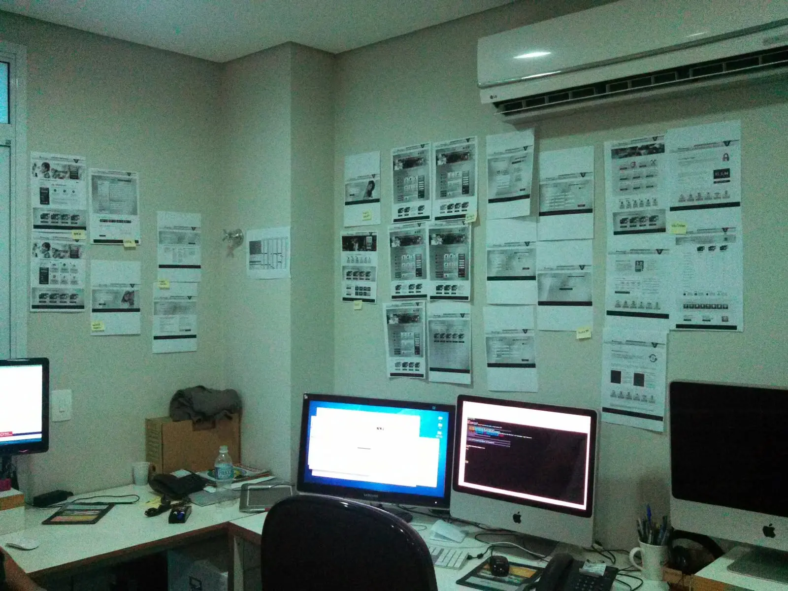 An office corner with web page layouts pinned across two walls above desks and computer screens