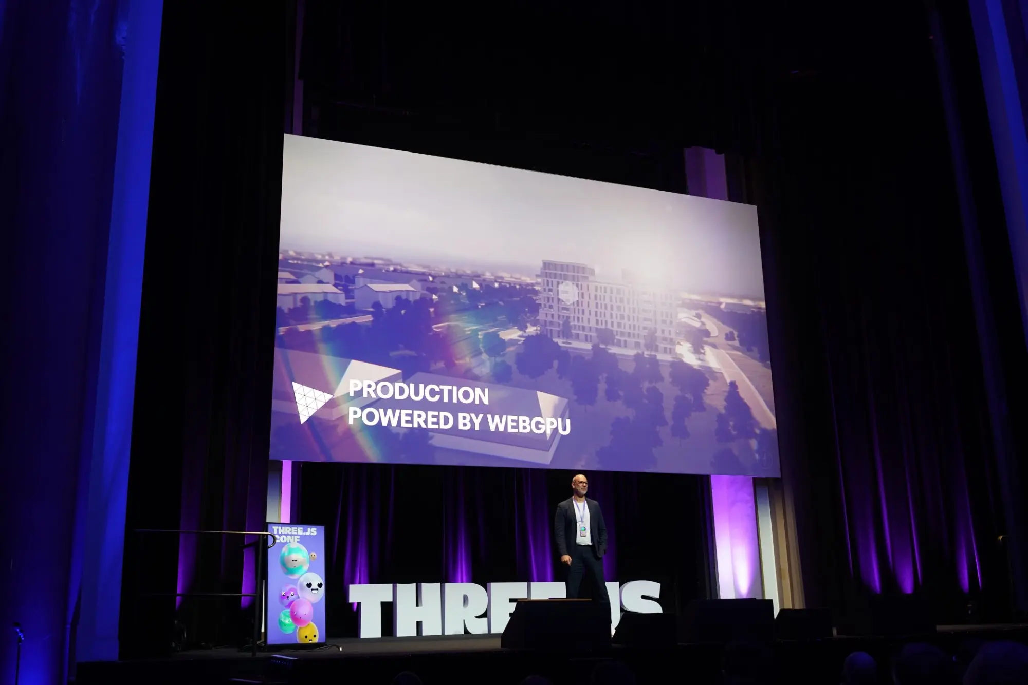 Anderson on stage at Three.js Conf Paris in front of a slide that reads Production Powered by WebGPU, over a rendered city scene