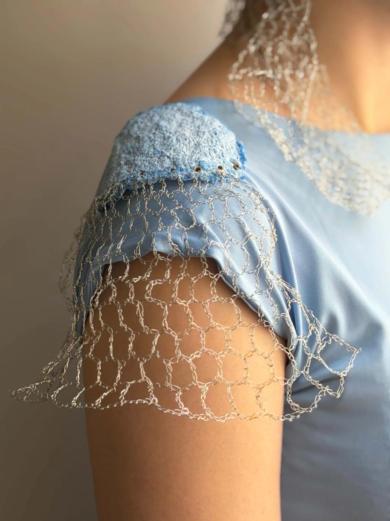Close-up of the shoulder piece: a blue textured panel with a row of small holes, and the crocheted wire mesh hanging from it over the sleeve and the upper arm
